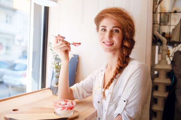 Smiling red haired woman sitting in cafe and eating dessert