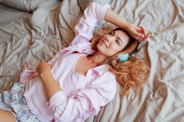 Smiling red head woman chilling on white bed in earphones in sunny morning after waking up.