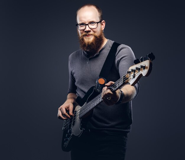 エレクトリックギターで遊んでいる灰色のTシャツを着た眼鏡をかけている笑顔の赤毛のひげを生やした男性ミュージシャン。灰色の背景に分離。