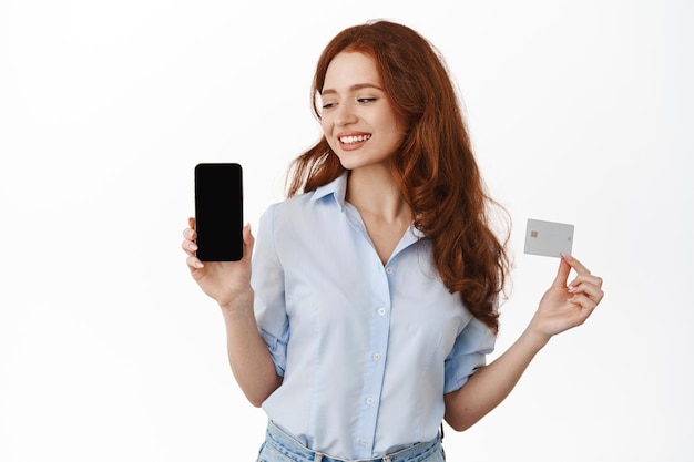 빈 전화 화면과 신용 카드를 보여주는 웃는 빨간 머리 사업가. 흰색 배경에 서서 스마트폰, 광고 은행 또는 상점에 만족하는 사무실 여성.