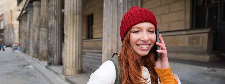 Free Photo smiling redhead female tourist talks on mobile phone and walks around city happy student in red hat