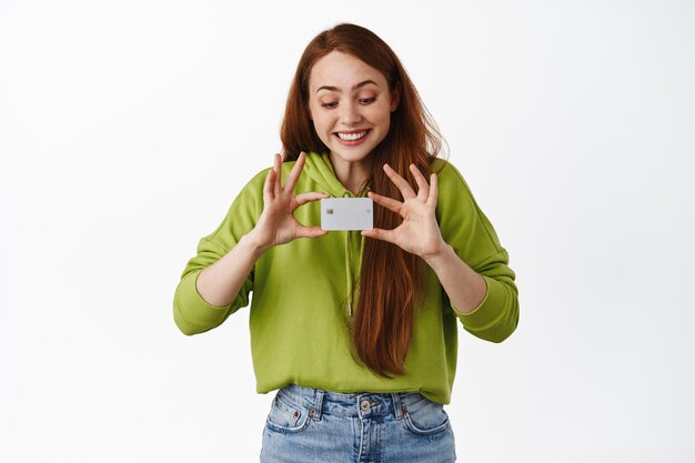 Smiling redhead girl looking at credit card, shopping contactless, making purchase, standing in casual outfit against white background. Copy space