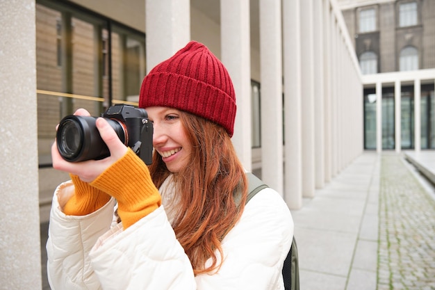 Улыбающаяся рыжая девушка-фотограф, фотографирующая в городе, делает фотографии на улице на профессиональном