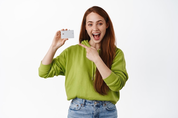 Smiling redhead girl pointing at credit card, recommend bank or shopping discounts, paying contactless, standing in casual clothes against white background