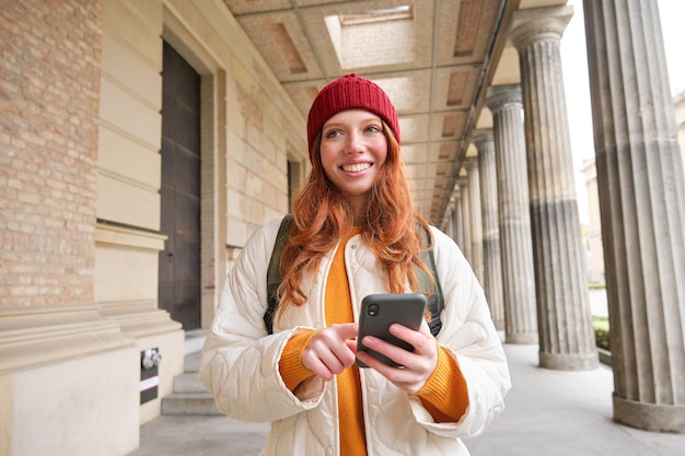 Smiling redhead girl in red hat looks at map on smartphone app checks her location searches for a ho