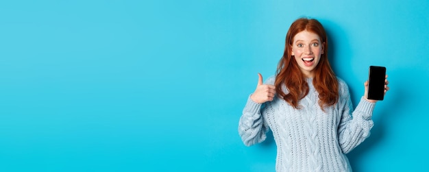 Free photo smiling redhead girl showing smartphone screen holding phone and demonstrating app making thumb up i
