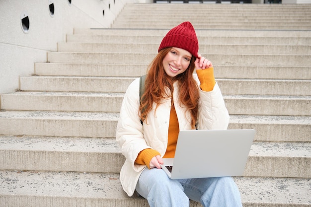 Foto gratuita giovane donna sorridente della ragazza della testarossa che digita sulla tastiera del computer portatile che si siede all'aperto sulle scale con il computer