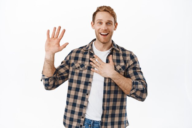Smiling redhead man holding one raised and another hand on heart, introduce himself, my name is nice to meet you gesture, saying hello, standing friendly and happy against white wall