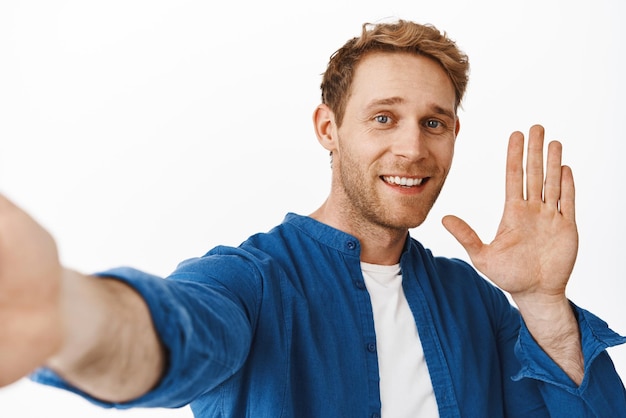 Бесплатное фото Улыбающийся рыжий мужчина машет рукой приветствие видеозвонка, держа камеру и делая селфи-блог или трансляцию в прямом эфире в приложении для смартфона, стоящем на белом фоне