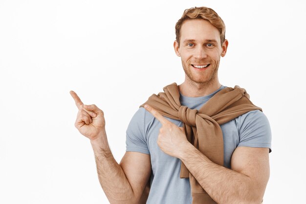 Smiling redhead man with muscles big biceps, pointing at upper left corner, showing advertisement on product on copy space, standing over white wall