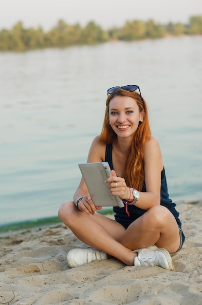 Улыбающаяся рыжая стройная женщина сидит на пляже и держит планшетный компьютер.