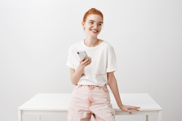 Smiling redhead woman lean on table, using mobile phone