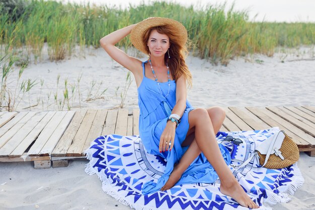 ビーチタオルの上に座って笑顔の赤毛の女性。完璧な日焼けボディ。青い服。風の強い髪。