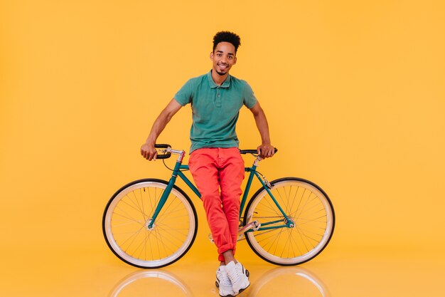 バイクの近くで喜んでポーズをとって黒髪の洗練された男を笑顔。緑の自転車で熱狂的なアフリカ人の屋内の肖像画。