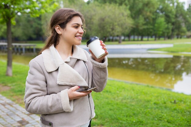 Улыбающаяся расслабленная девушка с телефоном пьет вкусный кофе