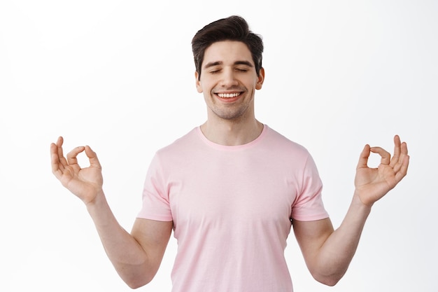 Foto gratuita sorridente uomo rilassato che medita, tenendosi per mano in un gesto zen om, sente pace e relax, respira tranquillo, in piedi su sfondo bianco