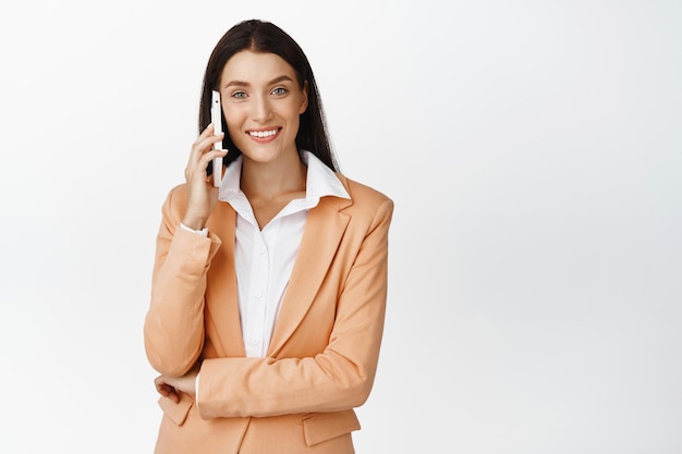 Улыбающаяся продавщица звонит клиенту Стильная деловая женщина разговаривает по мобильному телефону, стоя в костюме на белом фоне