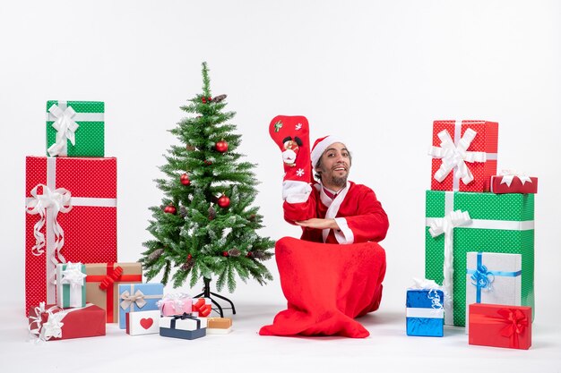 Smiling santa claus sitting on the ground and wear christmas sock near gifts and decorated new year tree on white background