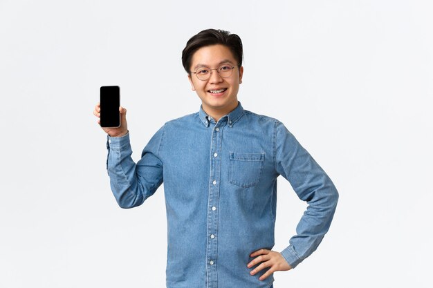 웃고 있는 만족스러운 아시아 남성 프리랜서, 스마트폰 화면을 보여주는 소규모 사업을 하는 기업가. 모바일 응용 프로그램을 사용하여 교정기와 안경을 쓴 남자, 흰색 배경