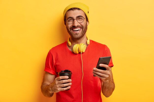 Smiling satisfied male wastes time in social networks, browses internet on mobile phone, drinks coffee from takeaway cup, has carefree joyful expression