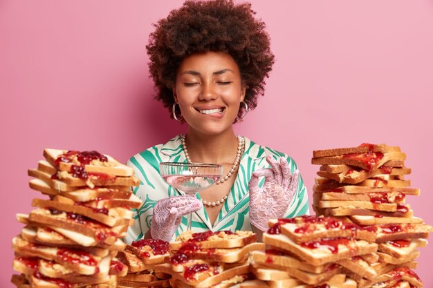 Smiling satisfied woman bites lips and closes eyes with pleasure, makes okay gesture, enjoys drinking cocktail, spends free time on banquet, stands near pile of bread sandwiches, celebrates birthday