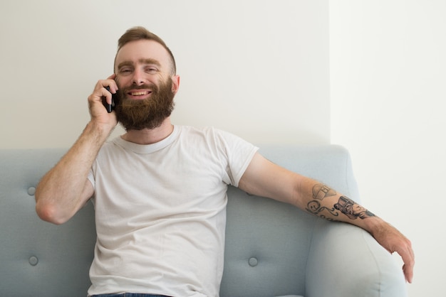 Giovane soddisfatto sorridente con la barba che per mezzo del telefono cellulare