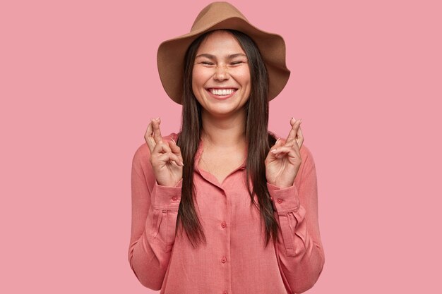 Smiling schoolgirl being full of wish, keeps fingers crossed, smiles broadly