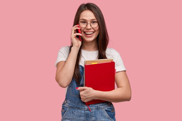 Smiling schoolgirl has phone conversation during break, holds modern cell phone, wears denim overalls, holds spiral notepad