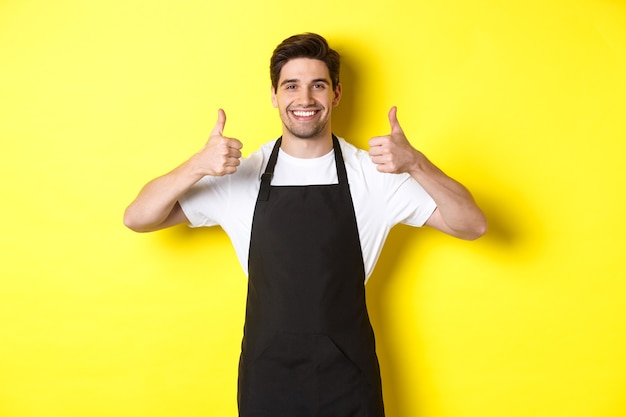 Smiling seller in black apron showing thumbs up, approve or like something, recommending cafe or store, yellow background.