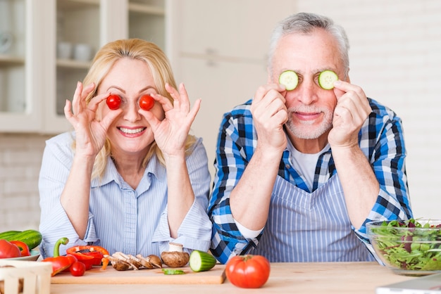 Foto gratuita coppie senior sorridenti che tengono i pomodori ciliegia e fette del cetriolo davanti ai loro occhi