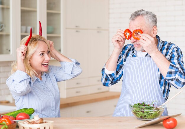 Smiling senior couple making fun with red chili peppers and bell peppers standing in the kitchen