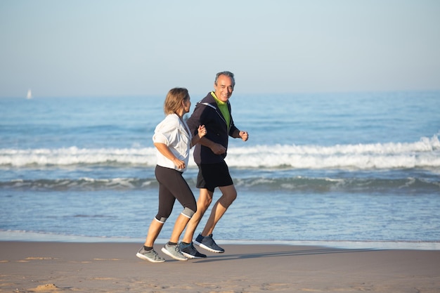 Smiling senior couple in sportswear jogging on sandy beach. Long shot of beautiful mature man and woman looking at each other, taking care of health on summer day. Sport, love concept