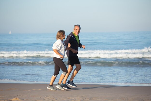砂浜でジョギングするスポーツウェアの老夫婦の笑顔。夏の日の健康管理をしながら、お互いを見つめ合う美しい成熟した男性と女性のロングショット。スポーツ、愛の概念