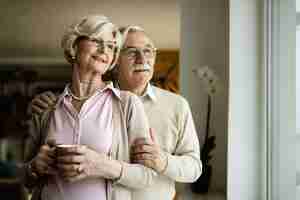 Free photo smiling senior couple standing embraced and looking through the window