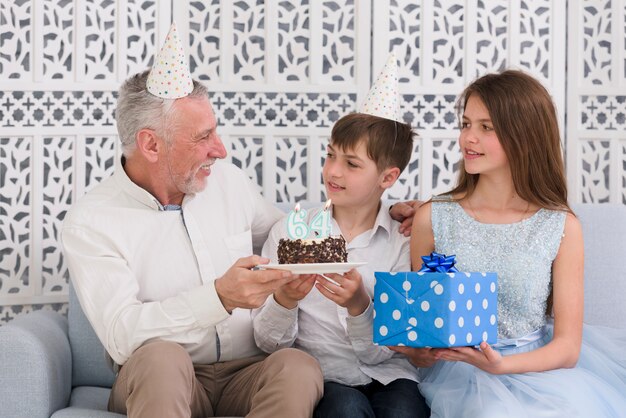 Smiling senior man looking at his grandchildren holding delicious birthday cake and gift box sitting on sofa