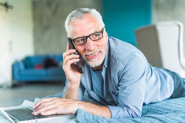 Smiling senior man lying over bed with laptop talking on cellphone