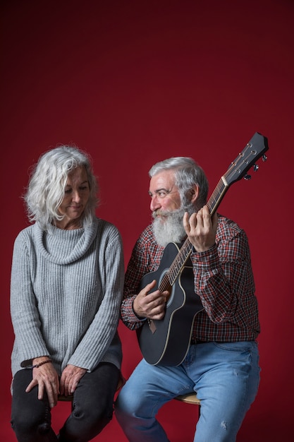Smiling senior man playing guitar for her wife sitting against red background
