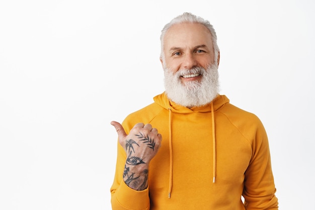 Smiling senior man with tattoos pointing left and looking happy, give advice, visit this page, click on link gesture, standing in orange hoodie against white wall