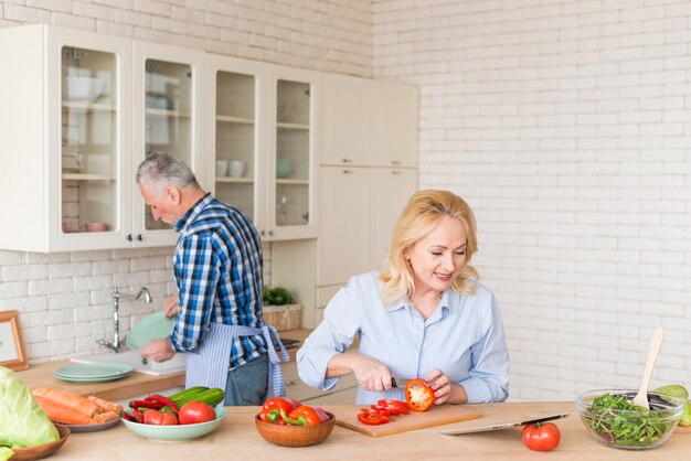 Усмехаясь старшая женщина режа красный болгарский перец с ножом смотря цифровую таблетку и его супруга моя посуду в кухонной раковине