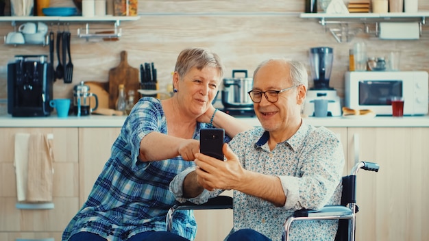 부엌에서 스마트폰을 사용하여 휠체어를 탄 노인 여성과 장애인 남편이 웃고 있습니다. 현대 통신 기술을 사용하는 마비된 노인 노인.