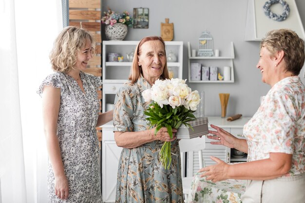 Smiling senior woman holding flower bouquet and gift standing with her daughter and grand daughter