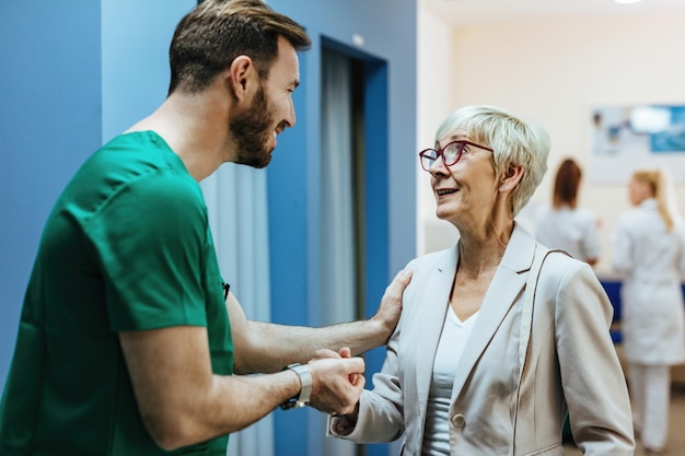 Donna anziana sorridente che si tiene per mano con un chirurgo mentre parla con il suo in un corridoio dell'ospedale