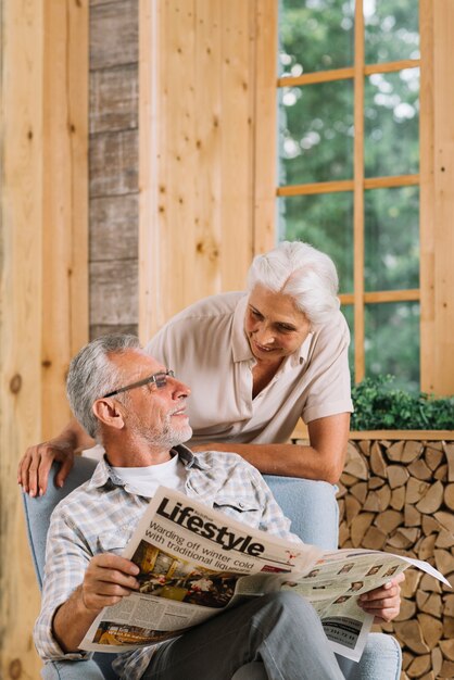 Smiling senior woman looking at her husband sitting on chair holding newspaper