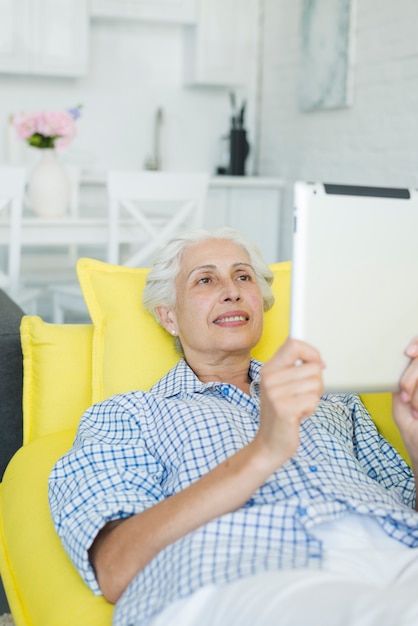 Smiling senior woman relaxing on yellow sofa looking at digital tablet