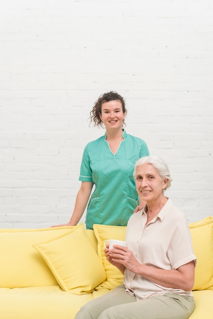 Smiling senior woman sitting on sofa holding coffee cup in front of nurse