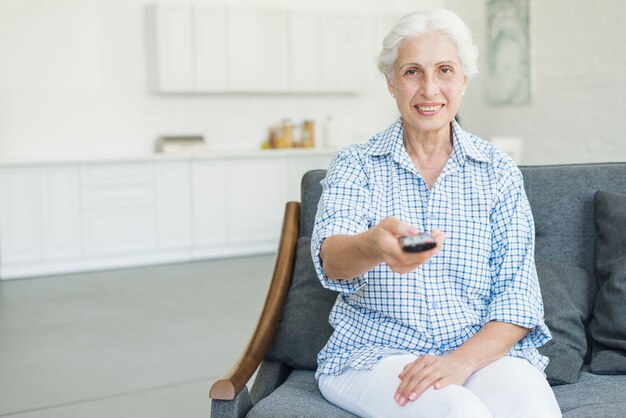 Улыбаясь старший женщина, сидя на диване, используя пульт дистанционного управления дома