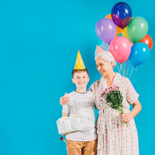 Smiling senior woman standing with grandson holding birthday gift on blue backdrop