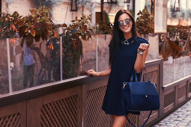 Smiling sexy brunette wearing stylish black dress in sunglasses, holds a black handbag, posing near a terrace cafe in the city.