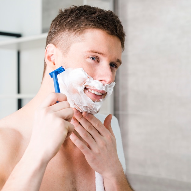 Free photo smiling shirtless young man shaving with blue razor