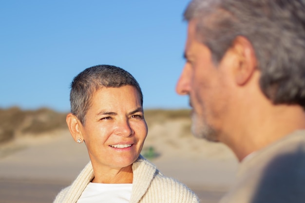 Smiling short-haired senior lady looking at her husband while couple spending time together on sunny evening outside. Grey-haired woman with brown eyes talking to bearded man. Relationship concept
